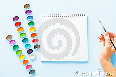 Creative flat lay of watercolor palettes, notebook, female hand holding paint brush. Artist workplace on a blue pastel background Stock Photo