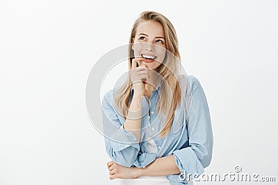 Creative good-looking female designer with blond hair, biting finger, looking up and smiling curiously while having Stock Photo