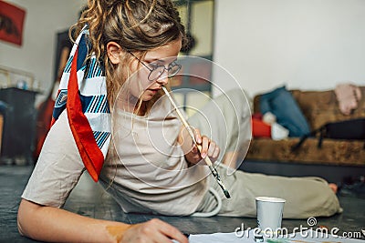 Creative young woman painting on paper in her studio. The female artist has colorful paint strokes on her face in a workshop Stock Photo