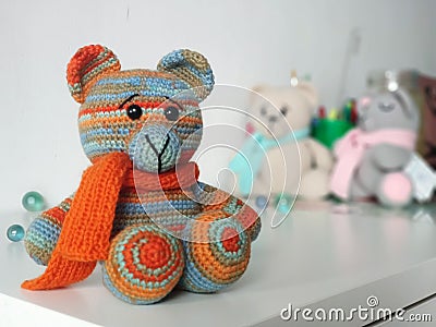 Crochet bear is orange-blue in scarf on background of other toys on light background. Knitted soft toy Teddy Stock Photo