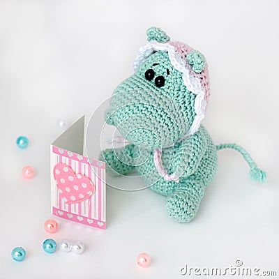 Crocheted amigurumi toy small turquoise hippo in a pink-white cap with a sad surprised look reads a book with a heart on cover Stock Photo
