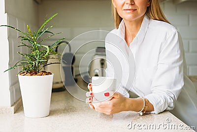 Crop photo of charming blonde woman with long fair hair in white shirt with morning cup of tea in hands on bright kitchen before Stock Photo