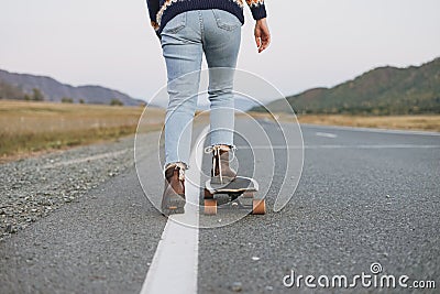 Crop photo of women`s legs in jeans on longboard on road with the mountains background Stock Photo