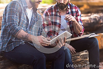 cropped image of bearded lumberjacks sitting on logs with axe and digital tablet Stock Photo