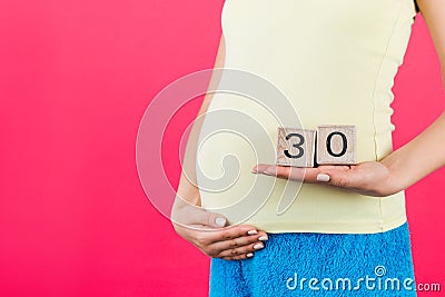 Cropped image of cubes with thirty weeks of pregnancy in pregnant woman`s hands wearing colorful home clothing at pink background Stock Photo