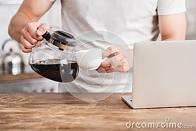 cropped image of man pouring coffee from coffee pot into cup near laptop Stock Photo
