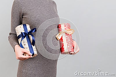 Cropped image of pregnant woman in gray dress holding two gift boxes at gray background. Is it a boy or a girl Waiting for twins. Stock Photo