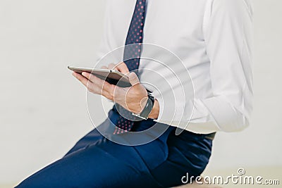 Cropped image of successful prosperous businessman in white shirt, black trousers, tie, holds modern tablet, checks notification, Stock Photo