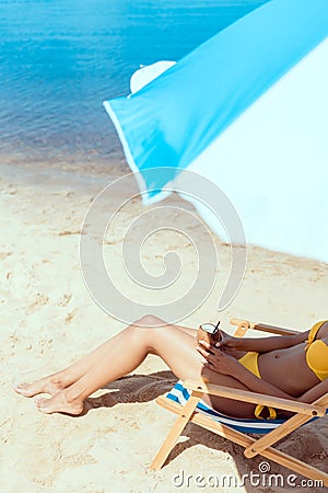 cropped image of woman in bikini laying on deck chair and holding cocktail in coconut shell under beach umbrella in front Stock Photo
