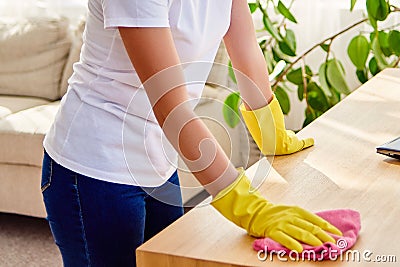 Cropped image of woman in white shirt and yellow protective rubber gloves cleaning at home and wiping dust on wooden tablel. Stock Photo