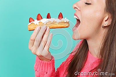 Cropped image of young woman in knitted pink sweater with closed eyes hold in hand, eating eclair cake isolated on blue Stock Photo