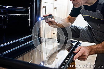 Cropped shot of aged repairman in uniform working, examining broken oven in the kitchen using flashlight. Repair service Stock Photo