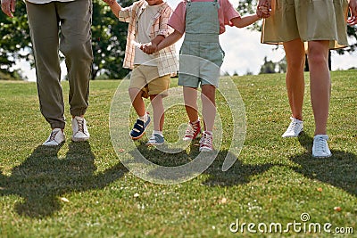 Cropped shot of legs of happy young family with two little kids holding hands, walking together in green summer park Stock Photo