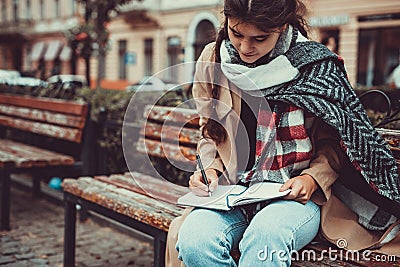 Cropped shot of lovely girl sitting on a wooden bench outside, writing a poem, looking lost in thoughts and dreams. Stock Photo