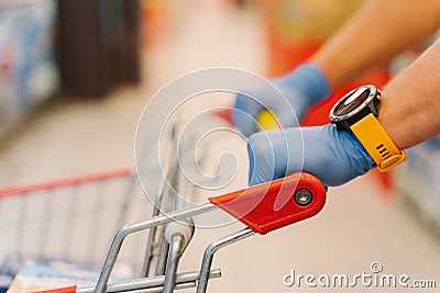 Cropped shot of mans hands in rubber gloves on handle of shopping cart against blurred supermarket background. Shopping, Stock Photo