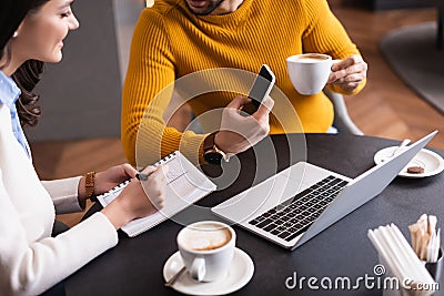 cropped view of freelancer showing smartphone to friend writing in notebook in restaurant. Stock Photo