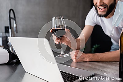 View of smiling man in medical mask holding glass of wine near laptop on kitchen worktop Stock Photo
