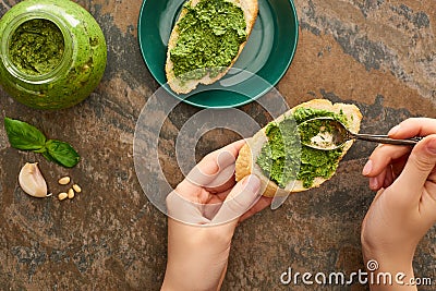 Cropped view of woman adding pesto sauce with spoon on baguette slice on stone surface Stock Photo