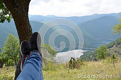 Crossed feet in sport shoes leaning on a tree and a blurred background with mountains and a lake Stock Photo