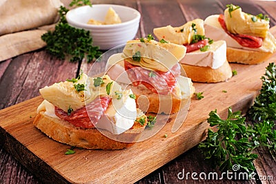 Crostini appetizers with brie cheese, salami and artichokes, close up on a serving board against a wood background Stock Photo