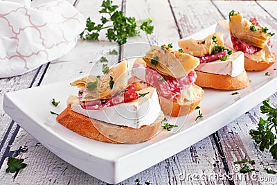 Crostini appetizers with brie cheese, salami and artichokes, close up on a plate against white wood Stock Photo