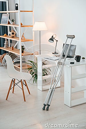 Crutches is leaning on the table in domestic room with laptop, chair and shelves Stock Photo