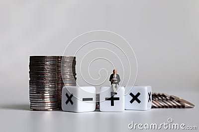 Cube arithmetic operator and a pile of coins. A miniature man sitting on a mathematical symbol cube. Stock Photo