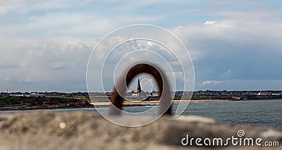 Cullercoats church overlooking Tynemouth Longsands beach, viewed through an old rusty Dock Ring on Tynemouth`s North Pier Stock Photo