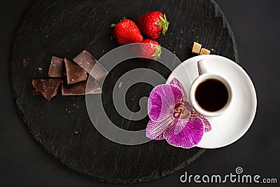 Cup of black coffee, brown sugar, strawberries, black chocolate on a round slate on a black background Stock Photo