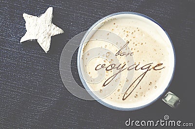 Cup of cappuccino coffee with foam in the form of words in French bon voyage on blue jeans, denim background. White wooden star. Stock Photo