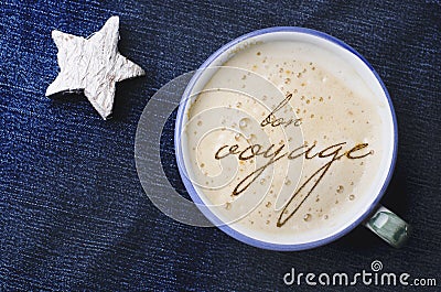 Cup of cappuccino coffee with foam in the form of words in French bon voyage on blue jeans, denim background. White wooden star. Stock Photo