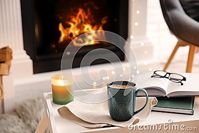Cup of coffee, burning candle and books on tray near fireplace indoors. Cozy atmosphere Stock Photo