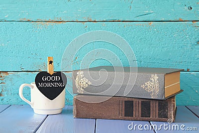 Cup of coffee with a little heart shape chalkboard with the phrase: GOOD MORNING next to stack of old books. Stock Photo