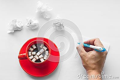 Cup of coffee on saucer with marshmallows, hand with pen writing on a blank sheet of paper and crumpled sheets of paper. Business. Stock Photo