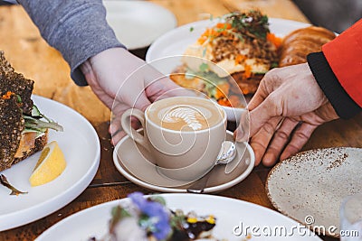 a cup of flat white coffee in cream ceramic cup with latte art on top, touched by two hands, surrounded with variety of food on Stock Photo