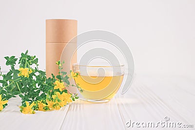 Cup of fresh herbal antioxidant tea, Hypericum flowers and blank paper packaging tube on white table. Alternative Stock Photo