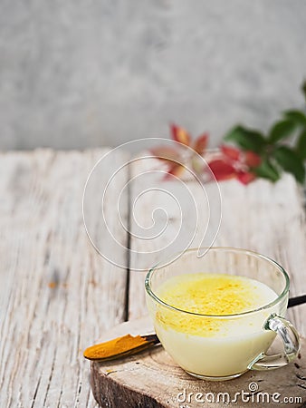 A cup with golden milk on a wooden stand. Near a wooden spoon with turmeric. Autumn leaves on a wooden background. Close-up. Copy Stock Photo