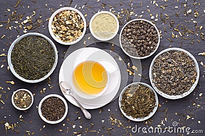 Cup of green tea and bowls of various dried tea leaves on dark, stone background Stock Photo