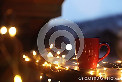 Cup of hot beverage on balcony railing decorated with Christmas lights, space for text. Winter Stock Photo
