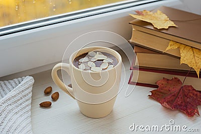 Cup of hot chocolate with almond and books on the windowsill. Window in a background. Fall time. Autumn composition. Home comfort Stock Photo