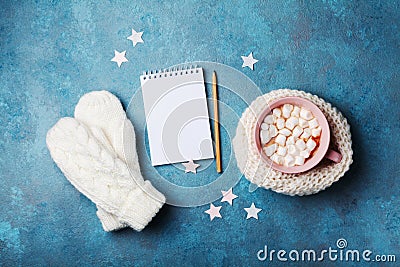 Cup of hot cocoa decorated knitted scarf with marshmallow, mittens and clean notebook on turquoise table top view. Flat lay style. Stock Photo