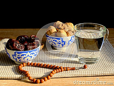 Cups with a traditional oriental pattern with dates and walnuts, a glass of clear water, stone rosary on a napkin during sunset Stock Photo