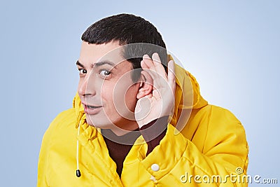 Curious brunet male model tries to overhear something, keeps hand on ear, listens gossips in next room, wears yellow anorak, isola Stock Photo