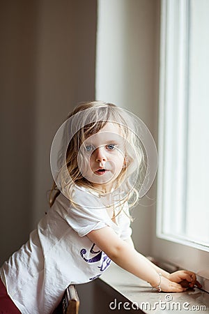 Curious cute little girl standing on a chair at the window, blonde curls Stock Photo