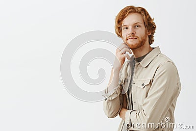 Curious friendly and relaxed kind redhead guy with beard and wavy hair holding hand on jaw standing half-turned at Stock Photo