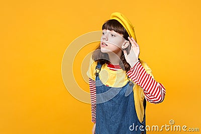 Curious girl teenager in french beret, denim sundress looking aside, eavesdrop with hearing gesture isolated on yellow Stock Photo