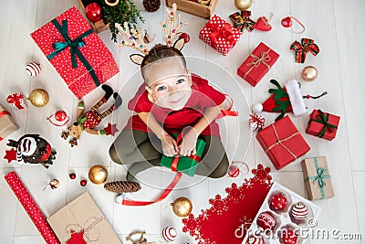 Curious girl wearing xmas costume reindeer antlers sitting on the floor, opening christmas present, top view. Stock Photo