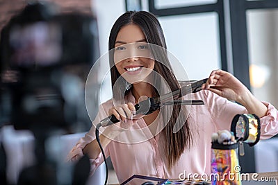 Beauty blogger smiling while curling hair and filming blog Stock Photo