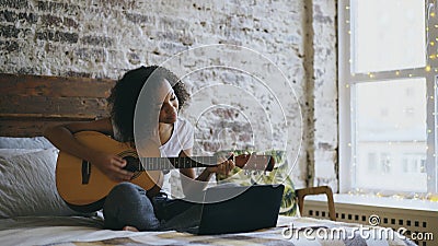 Curly african american teenager girl concentraing learning to play guitar using laptop computer sitting on bed at home Stock Photo