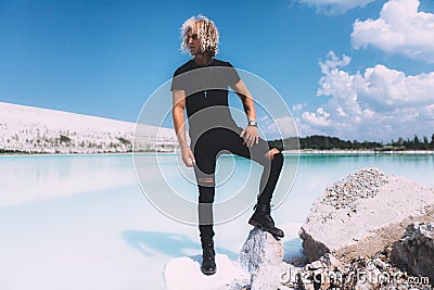 Curly blond man posing at the clear beach. Dressed all black. Awesome background, clear water and sky. Trendy, tropical and Stock Photo